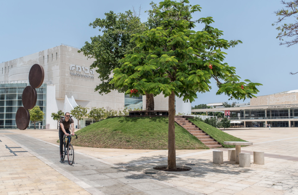 Habima square in daylight, showing two green trees. In the background, there is the Habima theater and tel aviv culture center. a man riding a bicycle